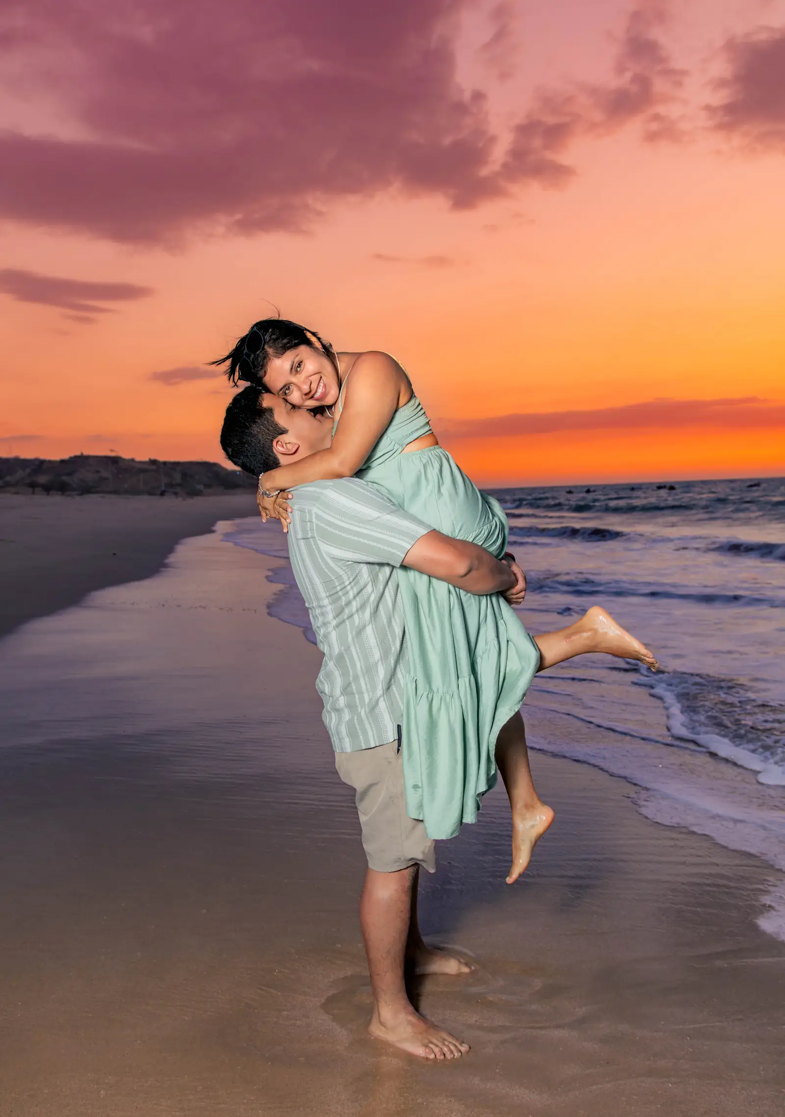 Pareja abrazada con la mujer en brazos de su pareja frente a un atardecer naranja en la playa